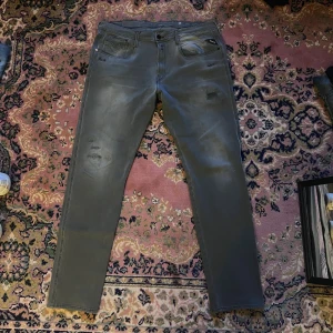 Replay Anbass grå jeans med slitningar 34/34 - Snygga Replay Anbass jeans i grått med diskreta slitningar på framsidan och klassiska fem fickor. Modellen har en slim passform och raka ben, med svarta detaljer på fickorna och logopatch bak. Perfekt för dig som gillar stilrena jeans med lite edge. Midja 46cm längd 107cm (33)