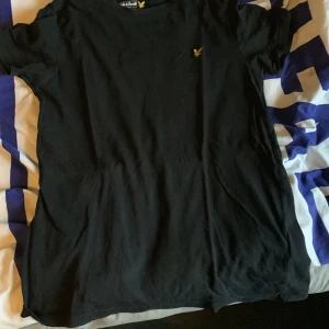 Svart t-shirt från Lyle & Scott - Svart t-shirt från Lyle & Scott med klassisk passform och gul broderad logga på bröstet. Enkel och stilren design, perfekt till jeans eller shorts. Tillverkad i mjuk bomull för skön känsla hela dagen.