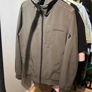 Grå vindjacka Peak Performance S - Snygg grön vindjacka från Peak Performance med huva och dragkedja framtill. Jackan har diskret logga på bröstet och ärmarna, samt praktiska fickor. Perfekt för dig som gillar stilren och sportig look. Använd få gånger!
