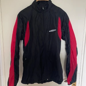 Svart och röd vindjacka från Newline - Sportig svart vindjacka från Newline med röda paneler längs ärmarna. Jackan har dragkedja framtill, snörning i nederkant och hög krage. Tillverkad i lätt polyester, perfekt för utomhusaktiviteter. Snygg och funktionell design med logga på bröstet. Passar M/S