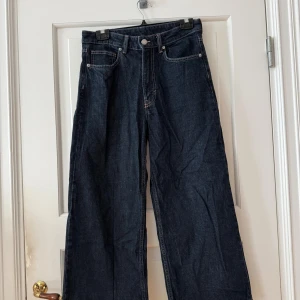 Mörkblå wide jeans från H&M, strl 40 - Snygga mörkblå jeans från H&M i modell wide med croppad längd och hög midja. Klassisk femficksdesign och raka, vida ben. Tillverkade i denim av 99% bomull och 1% elastan för lite stretch. Jeans från H&M. Storlek 40. Använda en gång. Hög midja, vida ben, ankellång. Ca 65 cm insida ben. Stretchiga. Mörk deminblå. Köpta för 299kr