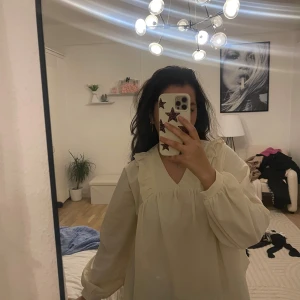 Ljus beige volangklänning med v-ringning - Säljer en söt och flowy ljus beige klänning med v-ringning och volangdetaljer vid axlarna. Klänningen har långa puffärmar med resår vid ärmslut och en avslappnad passform. Perfekt för dig som gillar romantiska och luftiga plagg.