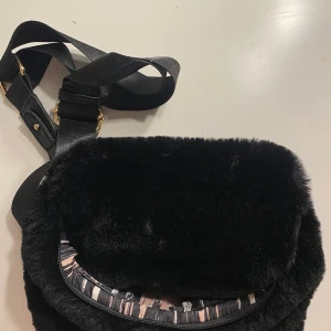 Svart axelväska med pälsdetaljer - Snygg svart axelväska  med mjuka pälsdetaljer och guldfärgade metalldetaljer. Väskan har ett justerbart band, dragkedja bak och ett coolt mönstrat innerfoder i rosa och svart. Perfekt för dig som vill sticka ut med en unik accessoar.