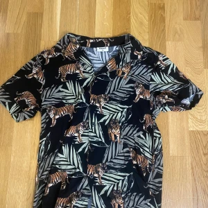 Svart skjorta med tiger- och bladmotiv - Cool svart skjorta från Kappahl med korta ärmar och knappar framtill. Skjortan har ett tropiskt mönster med tigrar och gröna blad som ger en riktigt unik vibe. Perfekt för dig som vill sticka ut med ett djuriskt print.