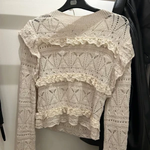 Beige stickad volangtröja - Supersöt beige stickad tröja med volangdetaljer och spets. Tröjan har ett genombrutet mönster och långa ärmar. Perfekt för dig som gillar romantisk och bohemisk stil. Snygg till både jeans och kjol!
