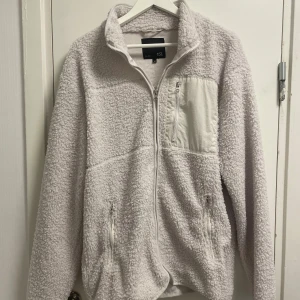 Vit teddyjacka från 157, storlek M - Mysig vit teddyjacka från 157 i storlek M. Jackan har hel dragkedja, två sidofickor med dragkedja och en bröstficka med dragkedja. Materialet är fluffigt fleece och jackan har hög krage för extra värme. Perfekt för kyliga dagar.