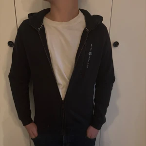 Svart hoodie från Sail Racing med dragkedja - Svart hoodie från Sail Racing med hel dragkedja framtill och broderad logga på bröstet. Tröjan har huva, lång ärm och en mjuk insida. Perfekt för dig som gillar en stilren och sportig look. Grisch Laidback Avicci Archive