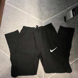 Svarta träningsbyxor från Nike - Svarta träningsbyxor från Nike med vit logga på ena benet. Byxorna har elastisk midja med snörning och är tillverkade i ett lätt syntetmaterial som passar perfekt för sport och aktivitet. Klassisk rak passform och diskret design.