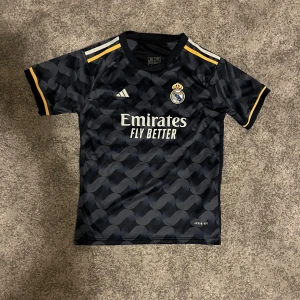 Real Madrid Vini Jr. Bortaställ - Snygg svart Real Madrid fotbollströja från Adidas med Vini Jr. och nummer 7 på ryggen. Tröjan har korta ärmar, gul och vit detalj på axlar och ärmslut, samt ett coolt abstrakt mönster. Broderad real Madrid märke och Adidas märke på bröstet.