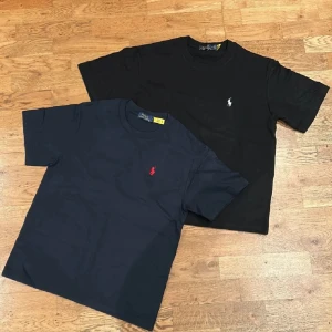 Svart och marinblå t-shirt Polo Ralph Lauren säljs tillsammans  - Två klassiska t-shirts från Polo Ralph Lauren i svart och marinblått. Båda har korta ärmar, rund hals och den ikoniska broderade loggan på bröstet – vit på den svarta och röd på den marinblå. Sköna i mjuk bomull och perfekt till jeans eller shorts.