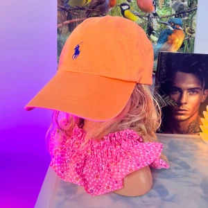 Orange keps från Polo Ralph Lauren - Snygg orange keps från Polo Ralph Lauren med klassisk blå broderad logga framtill och justerbart spänne bak. Kepsen är i bomull och har en avslappnad, sportig look som passar perfekt alla stilla kepsen är justerbar;)