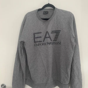 Grå sweatshirt från Emporio Armani EA7 - Säljer en stilren grå sweatshirt från Emporio Armani EA7 med stort logotryck framtill. Tröjan har rund halsringning och långa ärmar. Perfekt för en chill och snygg look. Materialet är mjukt och skönt, troligtvis bomullsmix. Har ett pytte litet hål på armen men inget man tänker på.