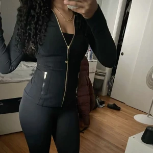 Svart slim träningsjacka från Lululemon - Snygg svart träningsjacka från Lululemon med guldiga dragkedjor och figurnära passform. Jackan har långa ärmar, två fickor med dragkedja och hög krage. Perfekt för dig som vill ha en sportig och stilren look.Inte använt mycket alls säljer den då jag vill köpa en annan färg.Den är i storlek S