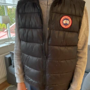 Svart dunväst från Canada Goose - Svart dunväst från Canada Goose med klassisk rund logotyp på bröstet. Västen har quiltad design och dragkedja framtill. Perfekt för lager-på-lager och har en stilren look med hög krage och diskreta sömmar. Passar dig som vill ha något varmt och snyggt. Färgerna, svart / väldigt mörkblå.