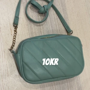 Grön quiltad axelväska med kedja - Snygg grön axelväska med quiltad design och guldfärgad kedjedetalj på axelremmen. Väskan har dragkedja upptill och en stilren, kompakt form som passar perfekt för dina viktigaste grejer. Enkel att matcha med olika outfits.