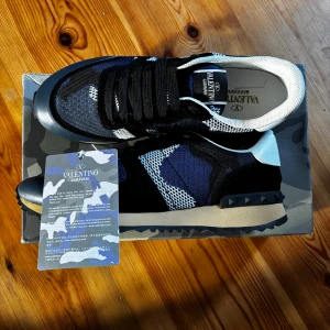 Valentino Rockrunner storlek 45 - Snygga Valentino Garavani Rockrunner sneakers i svart, blått och vitt med coolt kamouflagemönster. Skorna har snörning, platt sula med nitar och meshdetaljer för extra edge. Perfekta för dig som vill sticka ut med exklusiva och trendiga sneakers. Storlek 45.