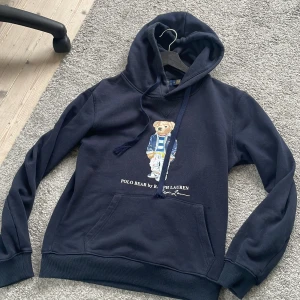 Mörkblå Polo Bear hoodie från Ralph Lauren - Säljer en mörkblå hoodie från Polo Ralph Lauren med ikoniska Polo Bear-trycket på bröstet. Tröjan har huva med snörning och en stor magficka. Tillverkad i mjuk bomull, perfekt för chill dagar. Klassisk och stilren streetwear-vibe. Den är i strlk M men passar både M och S. Den är i väldigt bra skick. MVH Arvid☺️