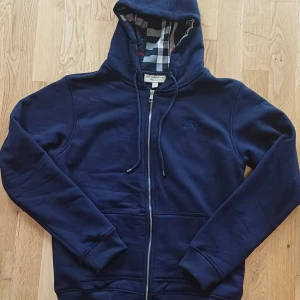 Marinblå Burberry hoodie med dragkedja - Snygg marinblå hoodie från Burberry i storlek L, tillverkad i 100% bomull. Klassisk dragkedja framtill, huva med snörning och ikoniskt rutigt foder i huvan och insidan. Storlek L men passar även som M.