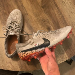 Nike fotbollsskor grå/orange med snörning - Säljer ett par Nike fotbollsskor i grå mocka med svart swoosh och cool orange sula med svartprickigt mönster. Skorna har snörning och rund tå, samt stickad detalj vid hälen för extra komfort. Perfekta för dig som vill sticka ut på planen.