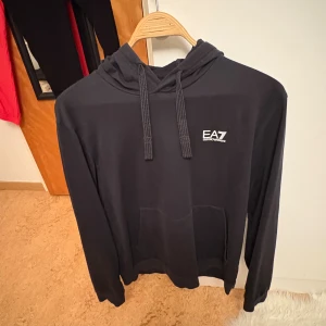 Svart hoodie från EA7 Emporio Armani - Svart hoodie från EA7 Emporio Armani med vit logga på bröstet. Tröjan har huva med snörning och en stor magficka framtill. Perfekt för en avslappnad och stilren look.