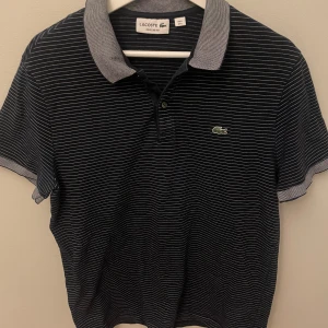 Randig blå Lacoste pikéskjorta - Snygg randig pikéskjorta från Lacoste i marinblått med tunna vita ränder. Klassisk krage i kontrasterande ljusare blå ton och ikoniska krokodilloggan broderad på bröstet. Regular fit och kortärmad, perfekt för en chill och stilren look.