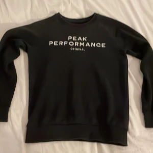 Svart sweatshirt från Peak Performance - Svart sweatshirt från Peak Performance med vit logga framtill. Klassisk rund halsringning och långa ärmar. Tröjan har en stilren och enkel design, perfekt för en avslappnad look. Tillverkad i mjukt material som känns skönt mot huden. Inga märken eller liknande. Säljer den för att den är för liten på mig