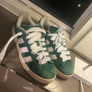 Adidas Gazelle gröna sneakers - Säljer ett par gröna Adidas Gazelle sneakers med klassiska vita ränder och logga på hälen. Skorna har vit snörning, platt sula och är tillverkade i mjuk mocka med vit innersida. Perfekt för dig som gillar retrostil och vill sticka ut med färg.