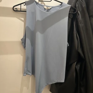 Ljusblå ärmlös linnebody med asymmetrisk kant - Säljer en ljusblå ärmlös linnebody med rund hals och cool asymmetrisk nederkant. Toppen har en clean och minimalistisk look, perfekt för dig som gillar stilrena plagg. Materialet känns mjukt och lätt mot huden.