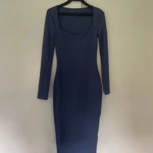 Marinblå ribbad långklänning - Snygg marinblå långklänning med ribbad struktur och långärmad design. Klänningen har en fyrkantig urringning och är figurnära med stretchigt material som sitter skönt mot kroppen. Perfekt för dig som gillar stilrena och enkla plagg med modern touch.