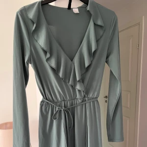 Ljusgrön omlottklänning med volang - Supersöt ljusgrön kort omlottklänning från H&M med långa ärmar, volangkant längs v-ringningen och knyt i midjan. Klänningen har en flowy passform och är gjord i mjuk polyester, perfekt för vår och sommar.