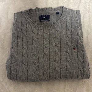 Grå kabelstickad tröja från Gant - Snygg grå kabelstickad tröja från Gant i 100% bomull. Perfekt för lager-på-lager och passar till allt från jeans till chinos.  Säljer en grå kabelstickad tröja från GANT med broderad logga på bröstet. Tröjan har rund halsringning och långa ärmar. Perfekt för dig som gillar klassisk och stilren look. Pris går alltid att diskutera. Orginalpris 999kr