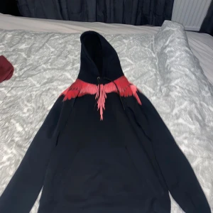 Marcelo Burlon svart hoodie med röd print - Svart hoodie från Marcelo Burlon med ikoniskt rött vingmönster över axlar och bröst. Tröjan har huva med snörning och känguruficka framtill. Tillverkad i 100% bomull för en skön och avslappnad känsla.