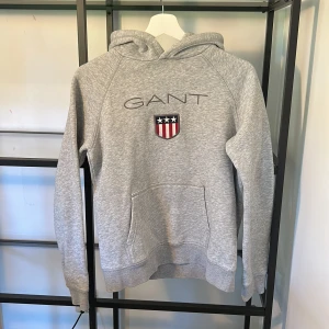 Grå hoodie från GANT med logga - Snygg grå hoodie från GANT med broderad logga och sköld på bröstet. Tröjan har huva, känguruficka och långa ärmar. Perfekt för dig som gillar klassisk och sportig stil. Mjuk och skön i bomullsmix.