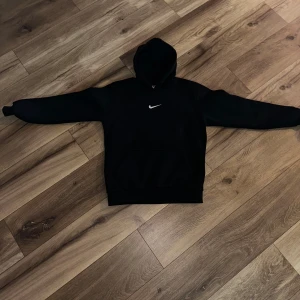 Svart Nike hoodie med vit logga - Svart hoodie från Nike med klassisk vit swoosh-logga på bröstet. Tröjan har huva, magficka och långa ärmar. Perfekt för en chill och sportig look. Mjuk insida och normal passform.