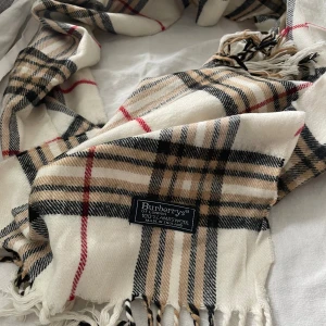 Rutig halsduk från Burberrys i ull - Klassisk rutig halsduk från Burberrys i 100% lammull. Beige, svart, vit och röd i det ikoniska rutmönstret med fransar i kanterna. Perfekt accessoar för att lyfta din outfit med en tidlös brittisk touch.