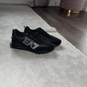 Svarta Emporio Armani sneakers - Snygga svarta sneakers från Emporio Armani med EA7-logga i vitt på sidan och Emporio Armani-tryck på hälen. Skorna har snörning, platt sula och rund tå. Materialmix av syntet och mocka ger en cool och modern look. Perfekta för dig som gillar stilrena och trendiga sneakers.