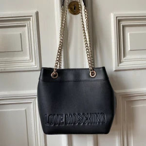 Love Moschino väska - Svart väska från Love Moschino. Kan bäras på axeln (som den hänger på bilden) eller crossbody. Fint använt skick. Metallen är lite skavd på ena sidan och skiftar i rosé, se tredje bilden. 