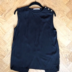  - Svart top från Acne studios med guldfärgade knappar på vänstra axeln och diagonal slits längs med ryggen.   Fint skick. Passar stl 34-36.   51% LYOCELL 49% MODAL   Tvättas i 30 grader fintvätt 