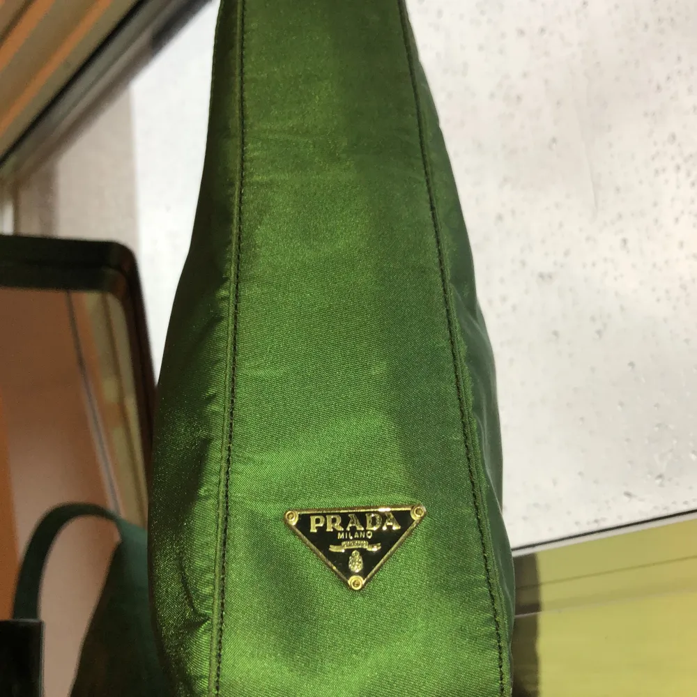 A Rare Prada shoulder bag in an asymmetrical green nylon style. This designer handbag features an asymmetrical body with a single shoulder strap and a Prada emblem near the base. The interior is tagged, “Prada Milano Made in Italy” beneath a zippered sidewall pocket in the signature Prada lining. A “63” tag marks the lower interior corner.  Condition  Good . Väskor.
