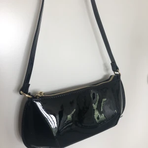 Liten svart väska  - Liten svart glansig väska, säljer då den tyvärr inte kommer till användning. Har ett kort axelband, en guldig dragkedja och en liten innerficka. Lite sliten på ena sidan (syns på bild 2) men annars är den i bra skick. Köparen står för frakt 🧚🏻‍♀️💘