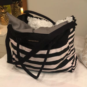 Väska - Jätte stor väska från Victoria’s Secret som rymmer hur mycket som helst. Två kuddar i på bilden för att demonstrera hur mycket den rymmer. Perfect för weekend trips eller liknande. Necessär i samma mönster medföljer. Kan även användas som iPad fodral. Kan placeras i framfickan av väskan. Färgerna är svart och ljusrosa. I mycket bra skick. Köparen står för frakt 📦 