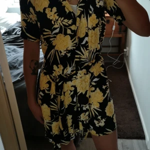 Klänning - Blommig sommarklänning. Snygg kombination i gult och svart. Luftig med knappar längs och med ett band att knyta med. 