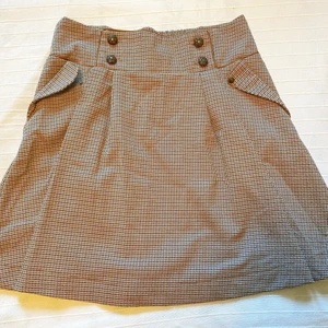 Smårutig mini-kjol med fickor - Kjol i härligt mjukt material, med stretch/gömd resår i midjan. Det smårutiga mönstret och fickorna gör den perfekt till skoldagar eller kontoret. Sitter som M men har taggen L på sig då den är köpt i Spanien. En dekorativ knapp saknas på framsidan.