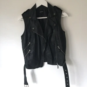  - Väst i bikermidell, svart veganskt läder och svart foder. Tre fickor med dragkedja, bälte med spänne längst ned. Många fina detaljer, t.ex axelklaffar.  Nyskick. Kan posta eller mötas upp i Stockholm. 