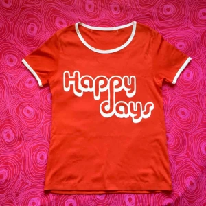  - 🍎 Röd t-shirt med vitt tryck där det står ’Happy days’. Obs! Det finns sprickor i texten 🍎